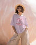 Person wearing a light purple t-shirt with text and graphics, standing against a soft pink background.